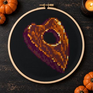 May include: A cross-stitch embroidery piece featuring a pixelated planchette design in shades of orange, brown, and dark purple, centred on a black fabric background and held within a circular wooden embroidery hoop frame.