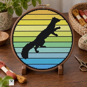 May include: A circular wooden embroidery hoop containing a cross-stitch pattern of a black ferret silhouette centered against a horizontal gradient background that transitions from yellow at the top to light green and blue at the bottom.