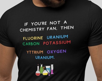 Op de afbeelding: Zwart T-shirt met de tekst "IF YOU'RE NOT A CHEMISTRY FAN, THEN FLUORINE URANIUM CARBON POTASSIUM YTTRIUM OXYGEN URANIUM." De tekst is in regenboogkleuren. Er zijn vijf bekers en kolven onder de tekst.