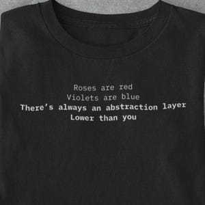 Op de afbeelding: Zwart t-shirt met de witte tekst "Roses are red, Violets are blue, There's always an abstraction layer, Lower than you". Het shirt ligt plat met een bril, smartphone, koffiebeker en andere items.