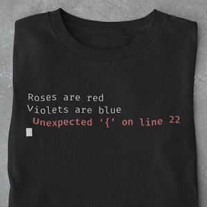 May include: A black t-shirt featuring the text "Roses are red, Violets are blue, Unexpected '{' on line 22" in red. The image includes a smartphone, glasses, a coffee cup, and a newspaper, all on a grey surface.
