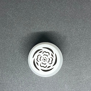 May include: A white, circular object with a floral design cut into the center. The object has a raised, rounded edge and sits on a gray surface. The floral design features a series of curved petals and a central cluster of small circles.