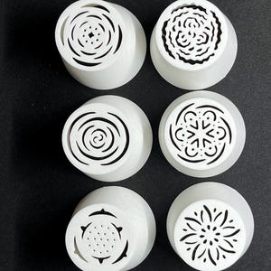 Puede incluir: Seis boquillas de decoración de pasteles de plástico blanco con diferentes diseños decorativos. Las boquillas tienen una base circular y una parte superior redonda con un diseño decorativo.