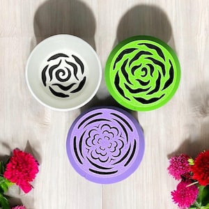 Könnte beinhalten: Drei runde, dekorative Lufterfrischer-Diffusoren mit floralen Designs. Die Diffusoren sind weiß, grün und lila. Der weiße Diffusor hat ein schwarzes Blumenmuster, der grüne Diffusor hat ein schwarzes Blumenmuster und der lila Diffusor hat ein lilafarbenes Blumenmuster.