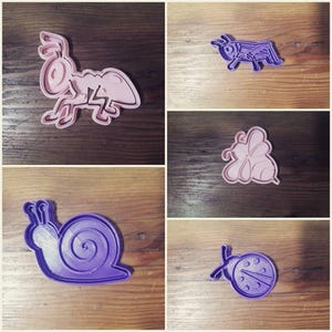 May include: A set of five cookie cutters shaped like insects and animals. The cutters are made of purple and pink plastic and feature a snail, ladybug, bee, cricket, and ant.