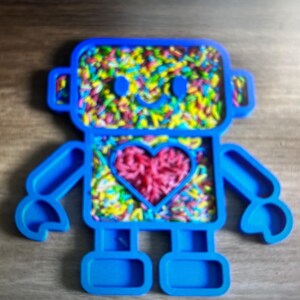 Könnte beinhalten: Ein blaues roboterförmiges Spielzeug, gefüllt mit bunten Streuseln. Der Roboter hat einen herzförmigen Ausschnitt in seiner Brust, der mit rosa Streuseln gefüllt ist. Das Gesicht des Roboters hat zwei blaue Augen und einen lächelnden Mund. Das Spielzeug ist für sensorisches Spiel konzipiert.