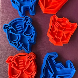 May include: Set of six cookie cutters featuring the Transformers Autobot and Decepticon logos. The cutters are made of blue and red plastic and are in the shape of the iconic robot faces.
