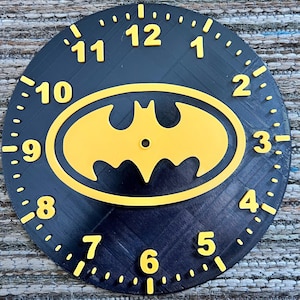 Pode incluir: Um mostrador de relógio redondo e preto com algarismos amarelos e o logotipo do Batman amarelo no centro. O mostrador do relógio tem uma superfície texturizada e é marcado com pequenos pontos ao redor do perímetro para indicar os minutos.