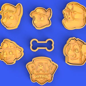 May include: A selection of yellow plastic cookie cutters featuring dog faces, a bone shape, and the Paw Patrol logo. The cutters are displayed against a blue backdrop.