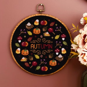 May include: A black embroidery hoop with a colourful autumn-themed cross-stitch design. The design features pumpkins, leaves, berries, and the word "AUTUMN" in a decorative font.