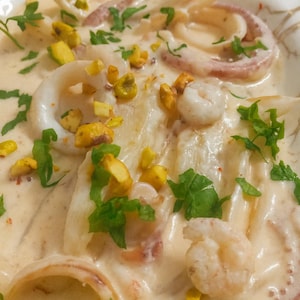 May include: A plate of creamy seafood, including calamari rings, prawns, and pasta, topped with chopped pistachios and fresh parsley. The dish is served in a white bowl, with a light-coloured sauce.