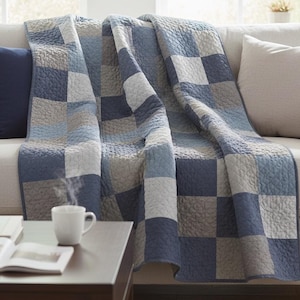 Pode incluir: Uma colcha xadrez azul, cinza e branca sobre um sofá bege. Uma almofada azul marinho repousa no sofá. Uma xícara de café branca está em uma mesa marrom escura, com um livro aberto.