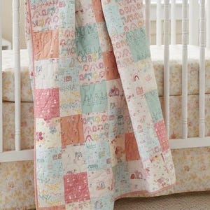 Könnte beinhalten: Eine Patchwork-Babydecke in Pastellfarben, drapiert über einem weißen Kinderbett. Die Decke zeigt Quadrate mit skurrilen Illustrationen von Häusern, Regenbogen und Tieren. Die Decke hat einen rosafarbenen Rand.