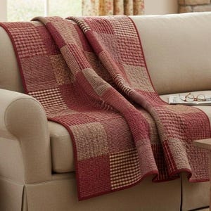 May include: A patchwork quilt featuring a repeating pattern of squares in shades of deep red, burgundy, and tan. The quilt displays various textures including small checkered prints, solid blocks, and woven patterns, draped over a beige sofa.