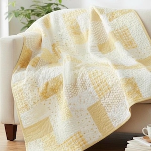 May include: A cream-colored quilt with a patchwork design in shades of yellow and white. The quilt is draped over a beige sofa, showcasing its textured patterns and stitching details. The quilt's dimensions are not visible.