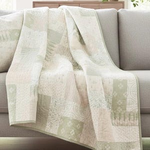 Puede incluir: Un edredón patchwork sobre un sofá gris. El edredón presenta una variedad de patrones en tonos de blanco, crema y verde salvia. El borde del edredón es de color verde salvia liso. El sofá está en una habitación bien iluminada.
