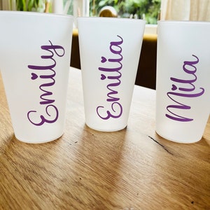 Könnte beinhalten: Drei weiße Becher mit lila Beschriftung. Die Becher sind mit "Emily", "Emilia" und "Mila" beschriftet.