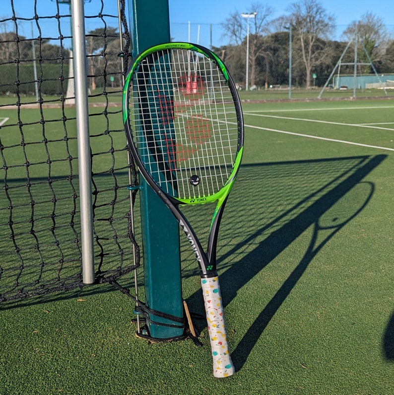 May include: A black and green tennis racket with a white grip featuring colorful cartoon designs. The racket is leaning against a green pole next to a tennis net on a green court. The racket's shadow is visible on the court.