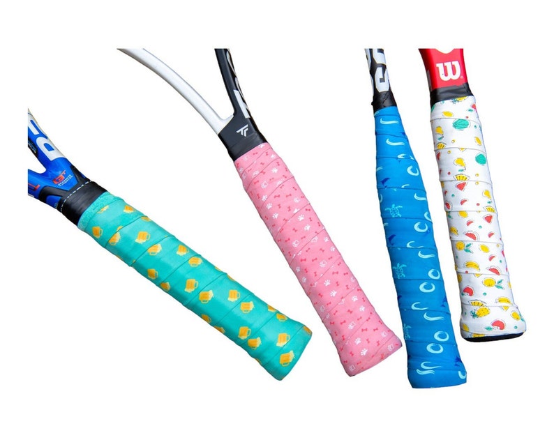May include: Four tennis racket grips in various colors. One is light green with yellow beer mugs, another is pink with small white paw prints, a third is blue with white turtles, and the last is white with colorful fruit.
