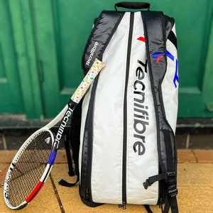 May include: A white and black tennis bag and racket set. The bag features the "Tecnifibre" logo in large black letters. The racket has a white, red, and blue design. The grip is wrapped in a colorful pattern.