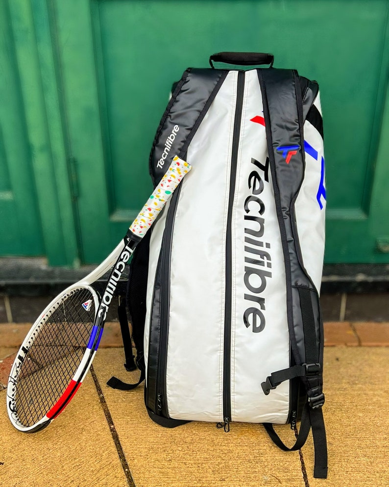 May include: A white and black tennis bag and racket set. The bag features the "Tecnifibre" logo in large black letters. The racket has a white, red, and blue design. The grip is wrapped in a colorful pattern.