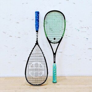 May include: Two squash rackets with black frames and stringing are displayed against a white wall. One racket has a blue grip with a pattern, the other has a turquoise grip with a yellow pattern. The rackets are for playing squash.