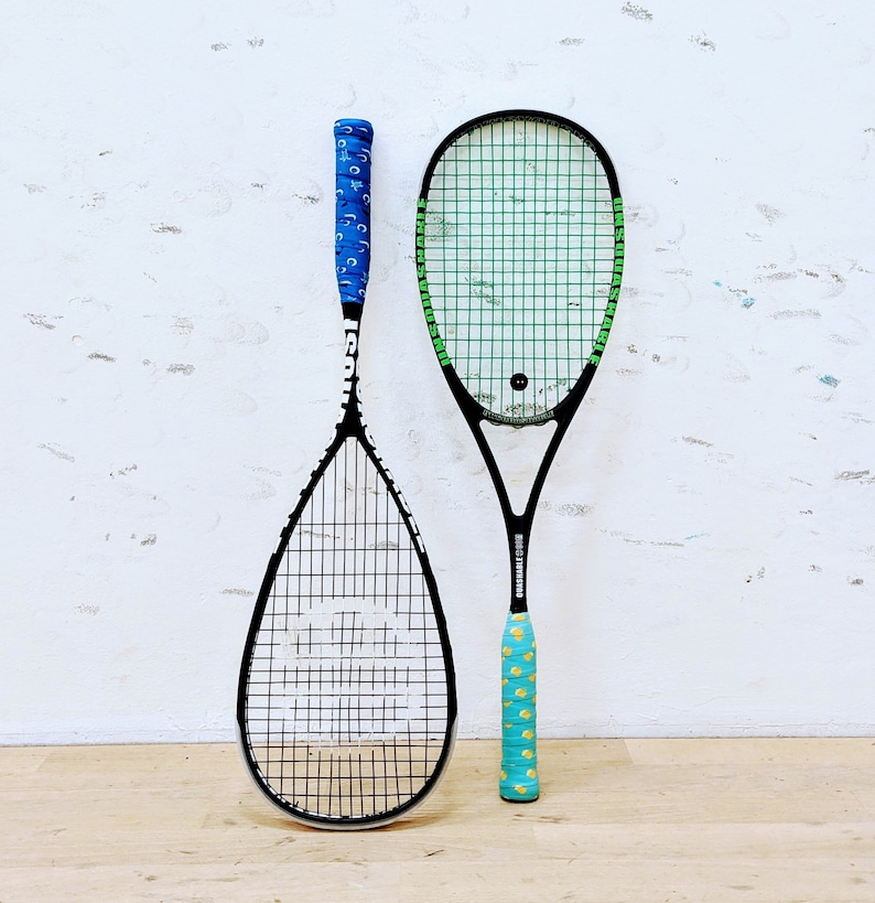 May include: Two squash rackets with black frames and stringing are displayed against a white wall. One racket has a blue grip with a pattern, the other has a turquoise grip with a yellow pattern. The rackets are for playing squash.