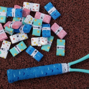 May include: A collection of colorful tennis grip tapes with various patterns, including fruits and geometric shapes, scattered on a textured surface. A tennis racket with a matching blue grip is also visible. The text "OLYMPIA GRIP" is printed on the grip tapes.