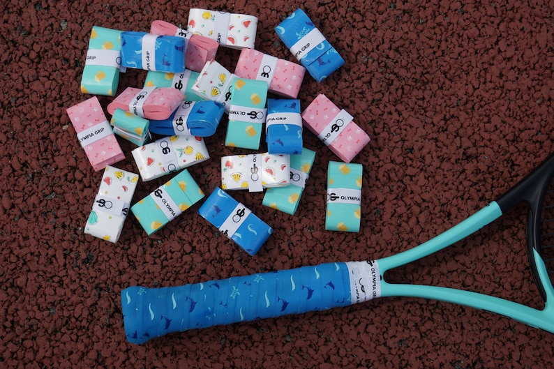 May include: A collection of colorful tennis grip tapes with various patterns, including fruits and geometric shapes, scattered on a textured surface. A tennis racket with a matching blue grip is also visible. The text "OLYMPIA GRIP" is printed on the grip tapes.