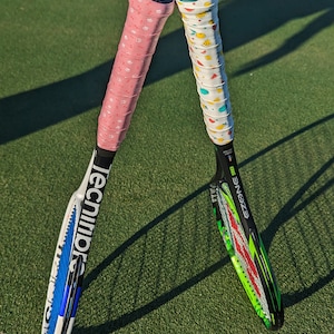 May include: Two tennis rackets with colorful grips rest on a green court. One racket has a pink grip with white star patterns, the other has a white grip with fruit designs. The rackets have black and white frames.