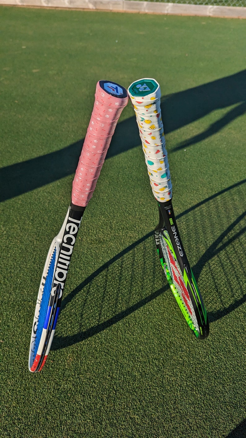 May include: Two tennis rackets with colorful grips rest on a green court. One racket has a pink grip with white star patterns, the other has a white grip with fruit designs. The rackets have black and white frames.