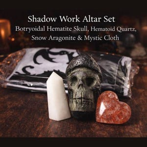 May include: A shadow work altar set including a Botryoidal Hematite skull, Hematoid Quartz heart, Snow Aragonite crystal, and a Mystic Cloth. The items are displayed on a dark wooden surface, with text at the top.