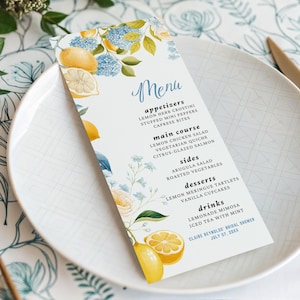 Può includere: Un biglietto del menu per un addio al nubilato con un design ad acquerello di limoni, ortensie blu e rose bianche. Il menu include antipasti, piatti principali, contorni, dessert e bevande. Il menu è su un piatto bianco.