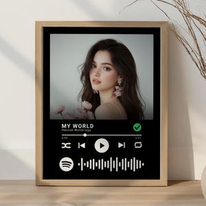 May include: Framed print featuring a woman's portrait above a black Spotify-style music player interface. The text "MY WORLD" and "Hannah Mondriaga" are visible. The frame is a light brown color.