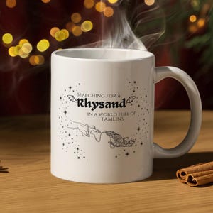 May include: White ceramic mug with a black graphic design that says "Searching for a Rhysand in a world full of Tamlins". The graphic features a hand reaching out to another hand with wings.