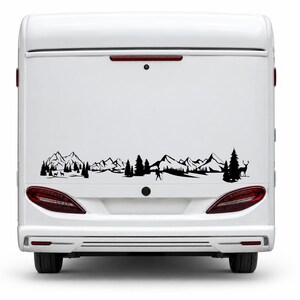 May include: A black vinyl decal applied to the rear of a white camper van. The design features a continuous landscape silhouette including jagged mountain peaks, dense pine forests, a lone hiker with a walking stick, and a standing deer.