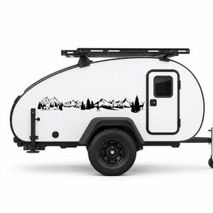 May include: A white teardrop camper trailer featuring a black roof rack and a black vinyl decal on its side. The decal depicts a detailed landscape scene with mountain peaks, evergreen trees, and silhouettes of deer in a forest setting.
