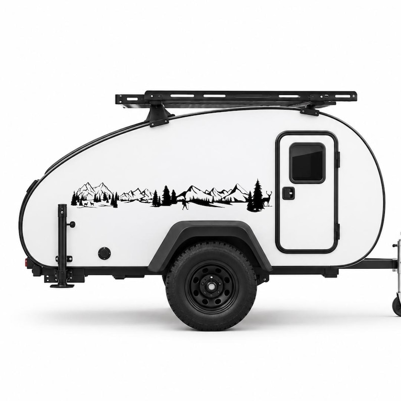 May include: A white teardrop camper trailer featuring a black roof rack and a black vinyl decal on its side. The decal depicts a detailed landscape scene with mountain peaks, evergreen trees, and silhouettes of deer in a forest setting.