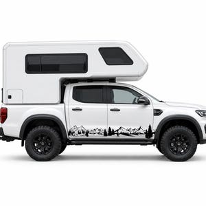 May include: A white pickup truck with a matching white camper shell features a black vinyl decal along the lower side panels. The decal depicts a detailed mountain range silhouette with evergreen trees, a small hiker, and wildlife.