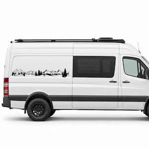 May include: A white van featuring a black vinyl decal on the side panel. The decal displays a detailed landscape illustration of jagged mountain peaks, dense pine trees, and a small deer silhouette, creating a nature-themed graphic design.