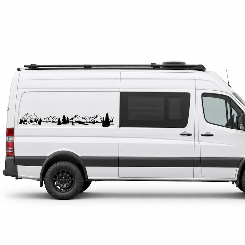 May include: A white van featuring a black vinyl decal on the side panel. The decal displays a detailed landscape illustration of jagged mountain peaks, dense pine trees, and a small deer silhouette, creating a nature-themed graphic design.
