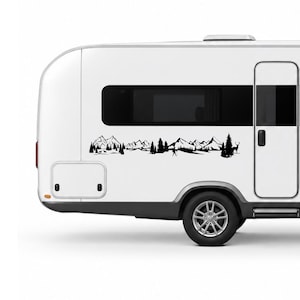 May include: A white camper trailer featuring a black vinyl decal of a mountain range landscape. The graphic includes silhouettes of jagged mountain peaks, dense pine trees, a small figure hiking with a pole, and a deer standing in the forest.