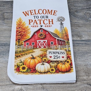 Könnte beinhalten: Ein weißes Waffelpiqué-Küchentuch mit einem herbstlichen Aufdruck, der eine rote Scheune, eine Windmühle sowie orangefarbene und weiße Kürbisse zeigt. Der Text lautet WELCOME TO OUR PATCH und PUMPKINS 25 cents, umrahmt von Sonnenblumen und Herbstlaub.
