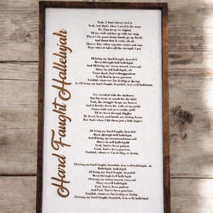 May include: A framed print with the words "Hard Faught Hallelujah" in a decorative font. The lyrics of the song are printed in a simple, easy-to-read font on a white background. The frame is a dark brown color.
