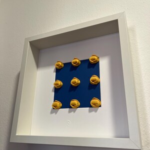 K&ouml;nnte beinhalten: Ein gerahmtes Kunstwerk mit neun gelben, pasta&auml;hnlichen Formen, die auf einem blauen Quadrat angeordnet sind. Das Kunstwerk ist vor einem wei&szlig;en Hintergrund in einem wei&szlig;en Rahmen platziert. Unten rechts befindet sich ein kleines, rundes Logo.