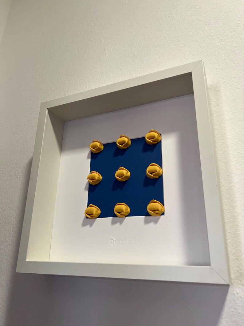K&ouml;nnte beinhalten: Ein gerahmtes Kunstwerk mit neun gelben, pasta&auml;hnlichen Formen, die auf einem blauen Quadrat angeordnet sind. Das Kunstwerk ist vor einem wei&szlig;en Hintergrund in einem wei&szlig;en Rahmen platziert. Unten rechts befindet sich ein kleines, rundes Logo.