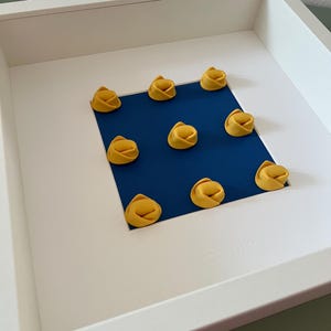 K&ouml;nnte beinhalten: Ein wei&szlig; gerahmtes Kunstwerk mit neun gelben Tortellini, die auf einem quadratischen blauen Papier angeordnet sind. Die Pasta befindet sich in einem wei&szlig;en Rahmen und schafft ein minimalistisches Kunstwerk zum Thema Essen.