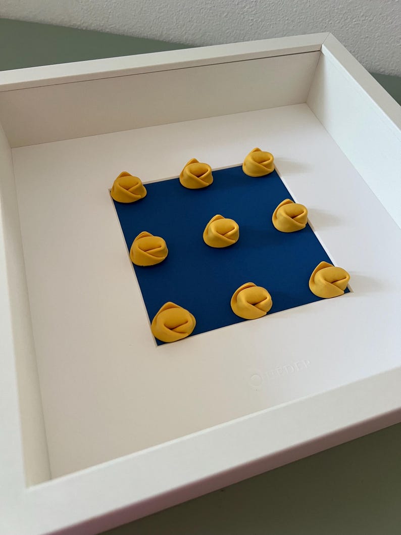 K&ouml;nnte beinhalten: Ein wei&szlig; gerahmtes Kunstwerk mit neun gelben Tortellini, die auf einem quadratischen blauen Papier angeordnet sind. Die Pasta befindet sich in einem wei&szlig;en Rahmen und schafft ein minimalistisches Kunstwerk zum Thema Essen.