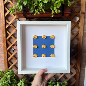 Può includere: Un'opera d'arte incorniciata in bianco con nove forme gialle a forma di semicerchio su sfondo blu. L'opera è tenuta in mano, con vegetazione e un traliccio di legno sullo sfondo.