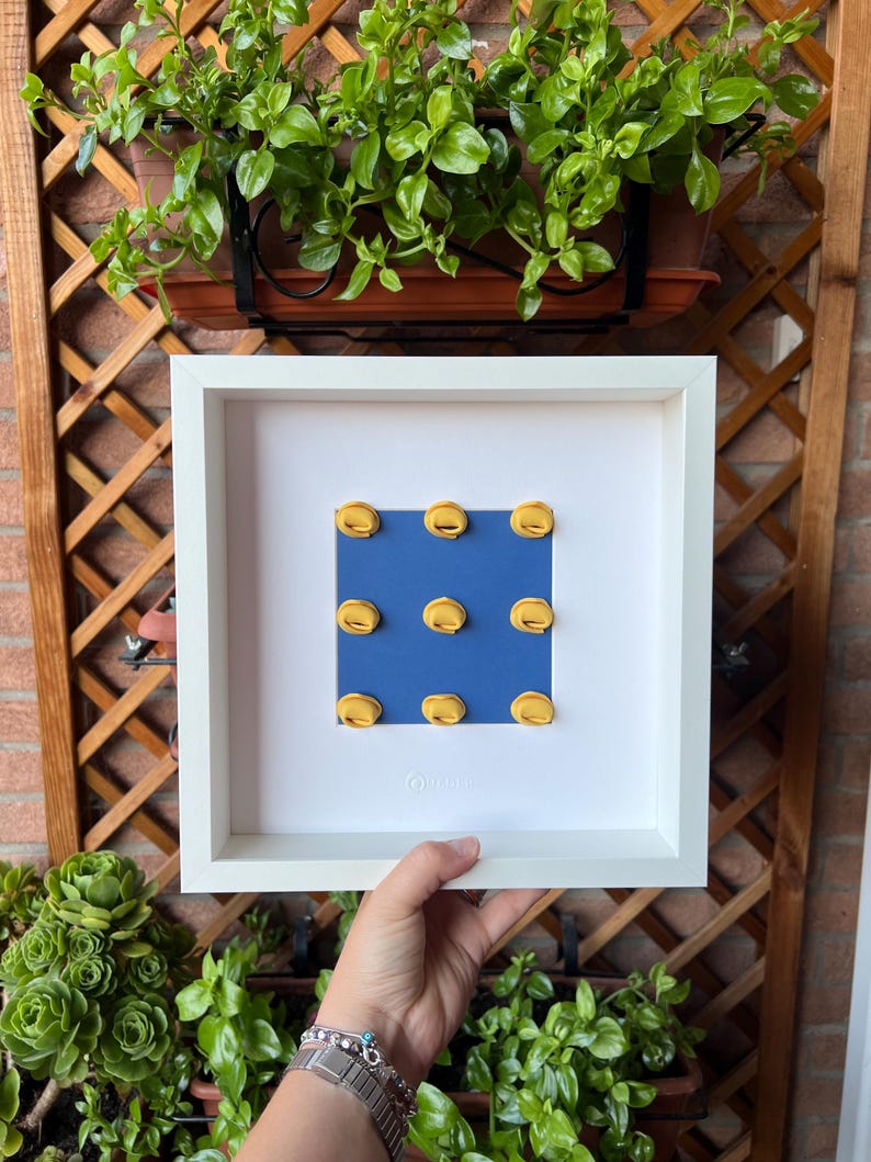 K&ouml;nnte beinhalten: Ein wei&szlig; gerahmtes Kunstwerk mit neun gelben, halbkreisf&ouml;rmigen Formen auf blauem Grund. Das Kunstwerk wird von einer Hand gehalten, mit Gr&uuml;n und einem Holzgitter im Hintergrund.